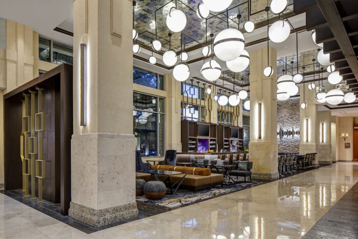 A modern hotel lobby with marble floors, tall columns, stylish seating areas, and a bar. Large round lights hang from the mirrored ceiling, and floor-to-ceiling windows let in natural light.