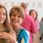 A smiling older woman helps a young woman try on earrings in a brightly lit store, with other women blurred in the background.