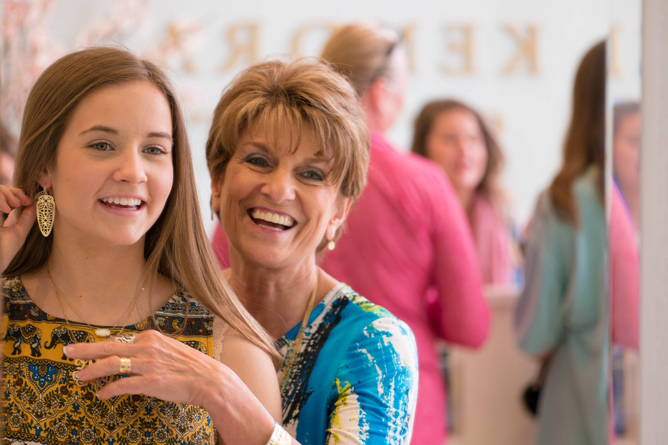 A smiling older woman helps a young woman try on earrings in a brightly lit store, with other women blurred in the background.