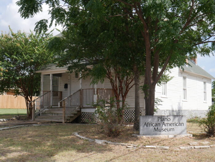 A white wooden house with a porch and trees in front, featuring a sign that reads “Plano African American Museum.” The house is surrounded by grass and a few bushes.