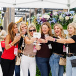 Six women stand together under a white canopy, smiling and raising glasses in a toast. They are surrounded by decorative flowers and appear to be enjoying a festive outdoor event.