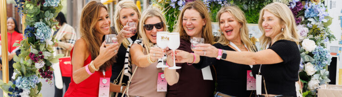 Six women stand together under a white canopy, smiling and raising glasses in a toast. They are surrounded by decorative flowers and appear to be enjoying a festive outdoor event.