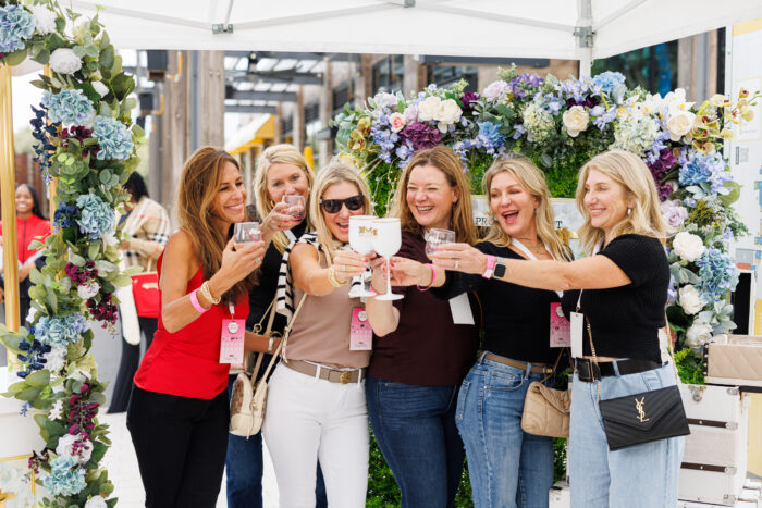 Six women stand together under a white canopy, smiling and raising glasses in a toast. They are surrounded by decorative flowers and appear to be enjoying a festive outdoor event.