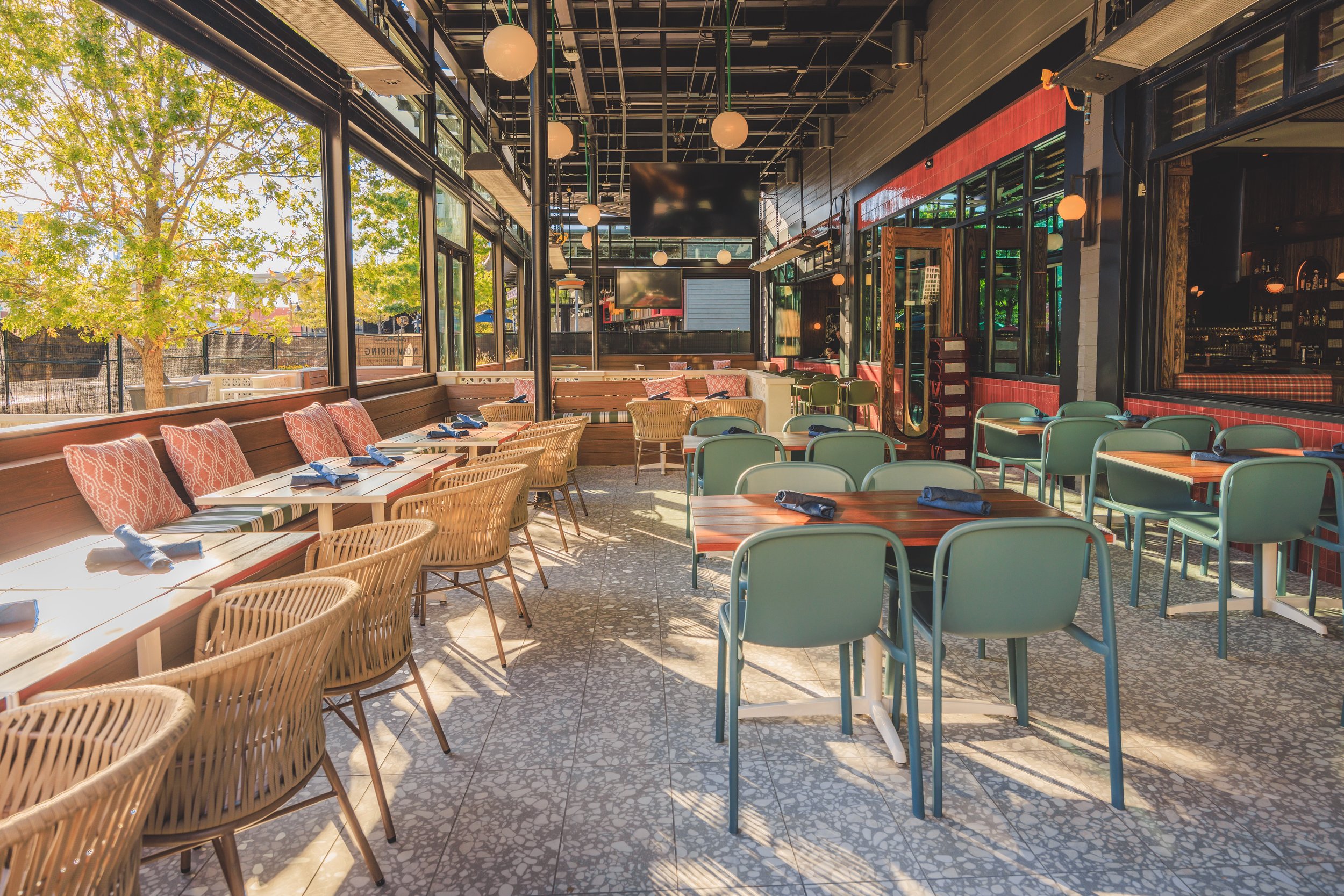 A sunlit restaurant patio with rattan chairs, green dining chairs, and wooden tables set for Group Experiences. Large windows overlook trees, while modern hanging lights enhance the inviting, airy atmosphere—perfect for memorable gatherings.