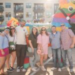 Eine Gruppe von sieben Erwachsenen steht lächelnd im Freien vor einer großen, bunten Piñata auf einem Fest und genießt das gemeinsame Erlebnis. Im Hintergrund sind Wohnhäuser und Verkaufsstände zu sehen.