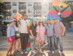 A group of seven adults stand smiling together outdoors in front of a colorful, large piñata decoration at a festival, enjoying shared group experiences. Apartment buildings and vendor tents are visible in the background.