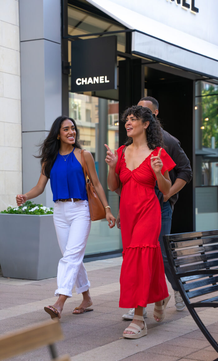 Two women walk and smile outside a Chanel store, one wearing a red dress and the other a blue top with white pants. A man walks slightly behind them. It appears to be a pleasant day for shopping.