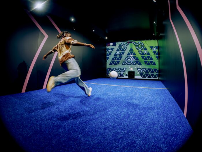 A person runs across a bright blue, indoor turf field toward a large soccer ball, surrounded by dark walls with geometric patterns and neon accents.