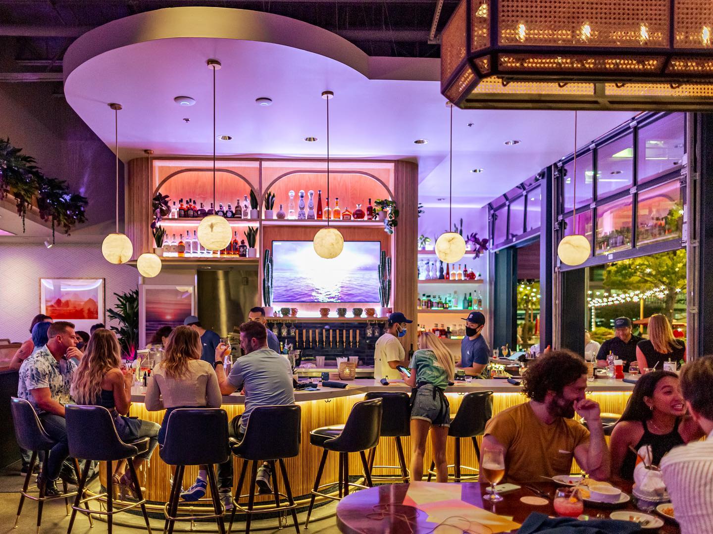 A modern bar with a glowing, curved counter and people enjoying group experiences on bar stools, chatting and drinking. Warm hanging lights and colorful neon accents create a lively, inviting atmosphere.