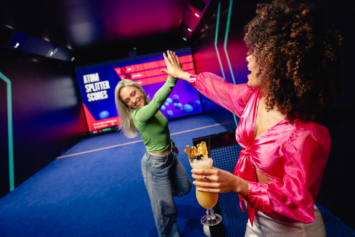 Two women celebrate with a high five in a neon-lit room. One, holding a tropical drink, wears a bright pink blouse; the other, in a green top and jeans, smiles joyfully. A digital scoreboard is visible in the background.