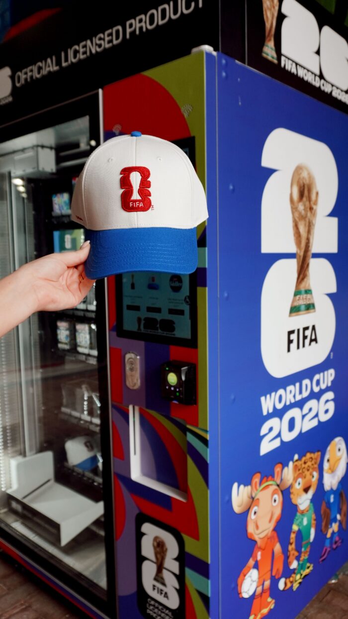 A hand holds a white and blue FIFA cap with the 2026 World Cup logo in front of a vending machine decorated with FIFA World Cup 2026 branding and mascots, celebrating the excitement of soccer in Plano.