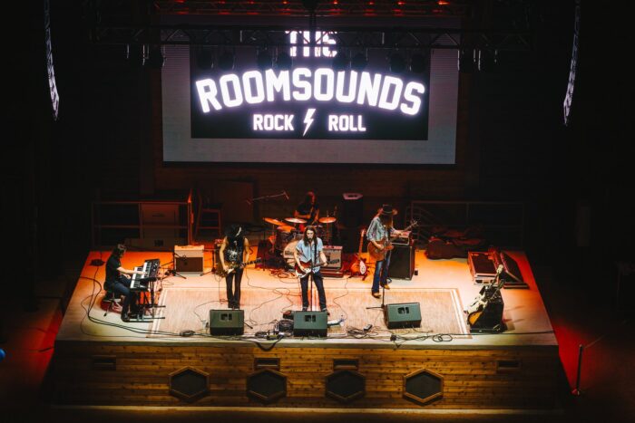 A band performs on stage under a large sign reading The Roomsounds Rock & Roll. Five musicians play instruments including guitar, keyboard, and drums, with stage lights highlighting their performance.