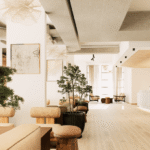 A spacious, modern lounge with light wood floors, neutral furniture, large windows, indoor plants, and abstract wall art, creating a bright and inviting atmosphere.