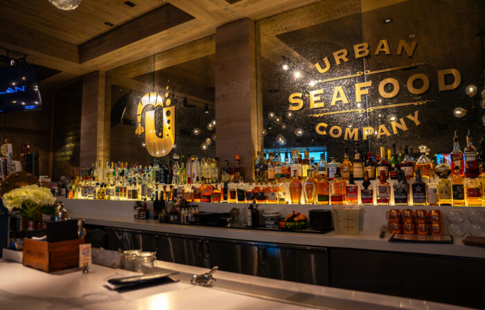 A well-lit bar with bottles of liquor lined up on shelves, a large mirror displaying the words Urban Seafood Company, and modern decor with warm lighting—perfect for memorable group experiences.