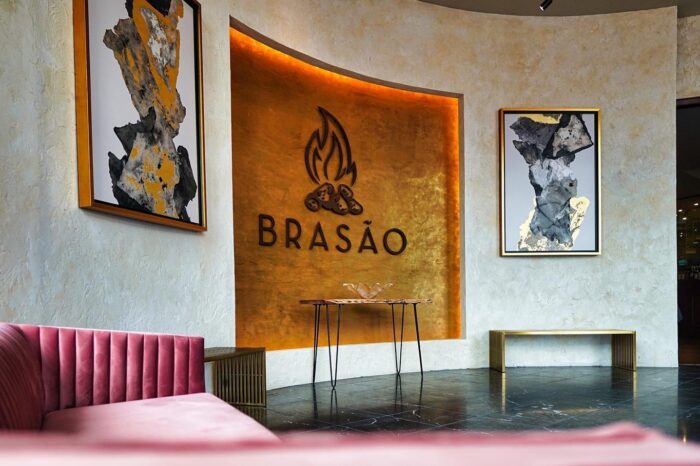 A modern restaurant interior with a gold textured wall featuring a fire logo and the word Brasão. Abstract art, a pink velvet bench, a gold bench, and a slim table accent the stylish space.
