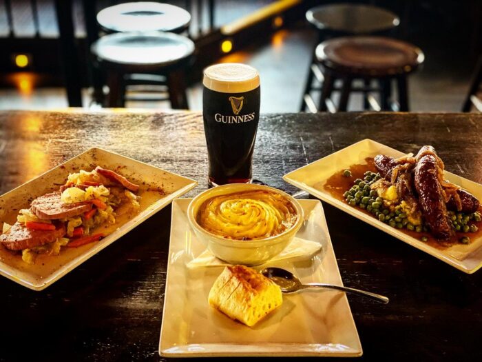 A pint of Guinness sits behind three plates of food—perfect for group experiences: sausage and cabbage, shepherd's pie with cornbread, and sausages with gravy, mashed potatoes, and peas—all on a wooden table in a cozy pub.