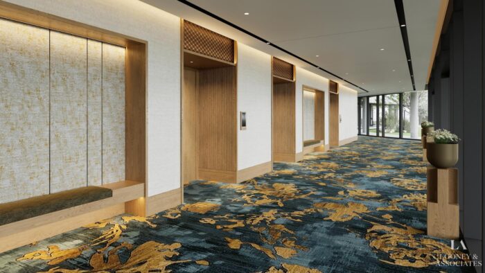 Modern hotel corridor with three wooden elevator doors, textured white walls, built-in bench seating, and a patterned blue and gold carpet. Floor-to-ceiling windows at the end let in natural light.