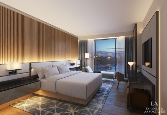 Modern hotel room with a large bed, wooden accent wall, soft lighting, a rug, armchair by a window overlooking an evening cityscape, and a desk with a TV on the opposite wall. Looney & Associates logo is in the corner.