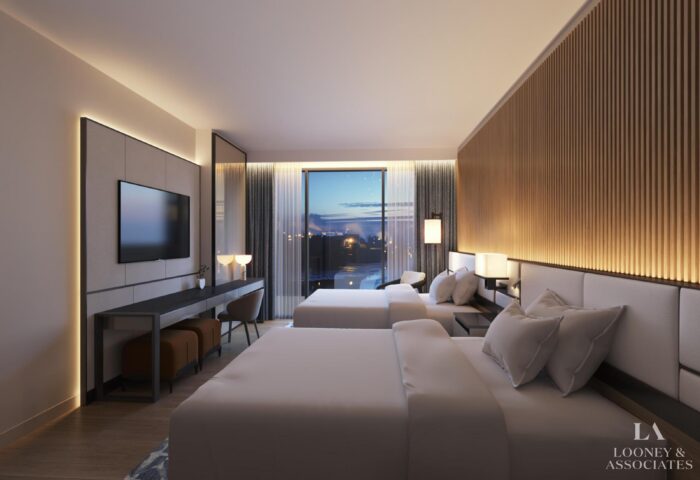 Modern hotel room with two beds, a large wall-mounted TV, a long desk with chairs, and floor-to-ceiling windows overlooking a pool and sunset, featuring warm lighting and wooden accent walls.