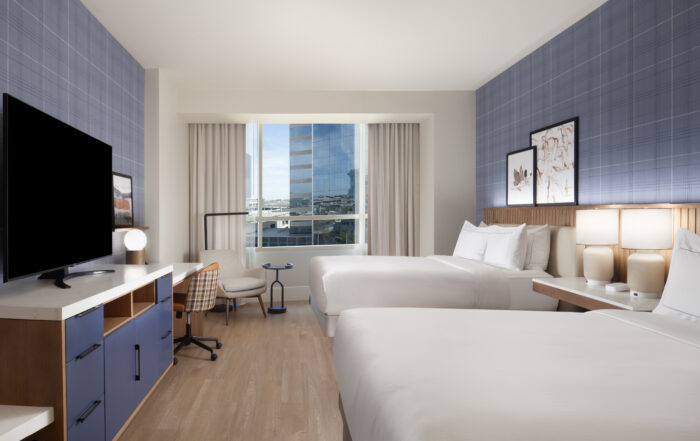 Modern hotel room with two large beds, a wall-mounted TV, a work desk with a chair, and large window with a city view. The room features blue accent walls, wooden flooring, and contemporary decor.