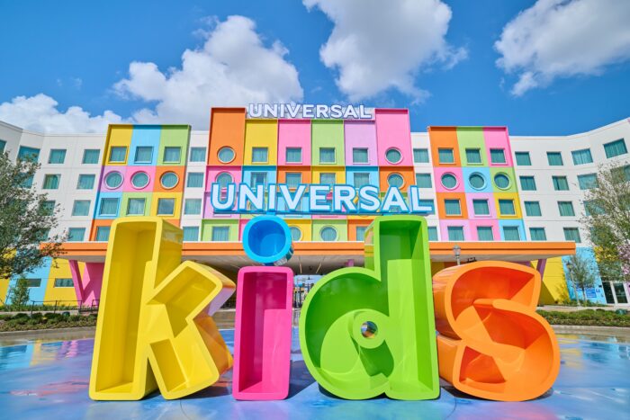 Colorful UNIVERSAL Kids sign in front of a vibrant hotel with square and circular windows, set against a blue sky with clouds.