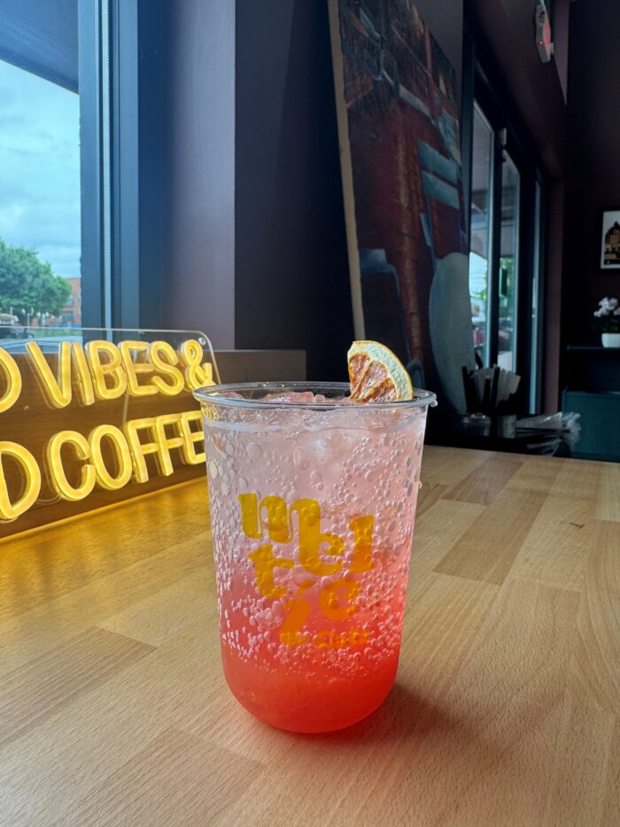 A clear plastic cup with a pink and red sparkling drink, topped with a dried citrus slice, sits on a wooden counter. In the background, a neon sign reads GOOD VIBES & GOOD COFFEE next to large windows.