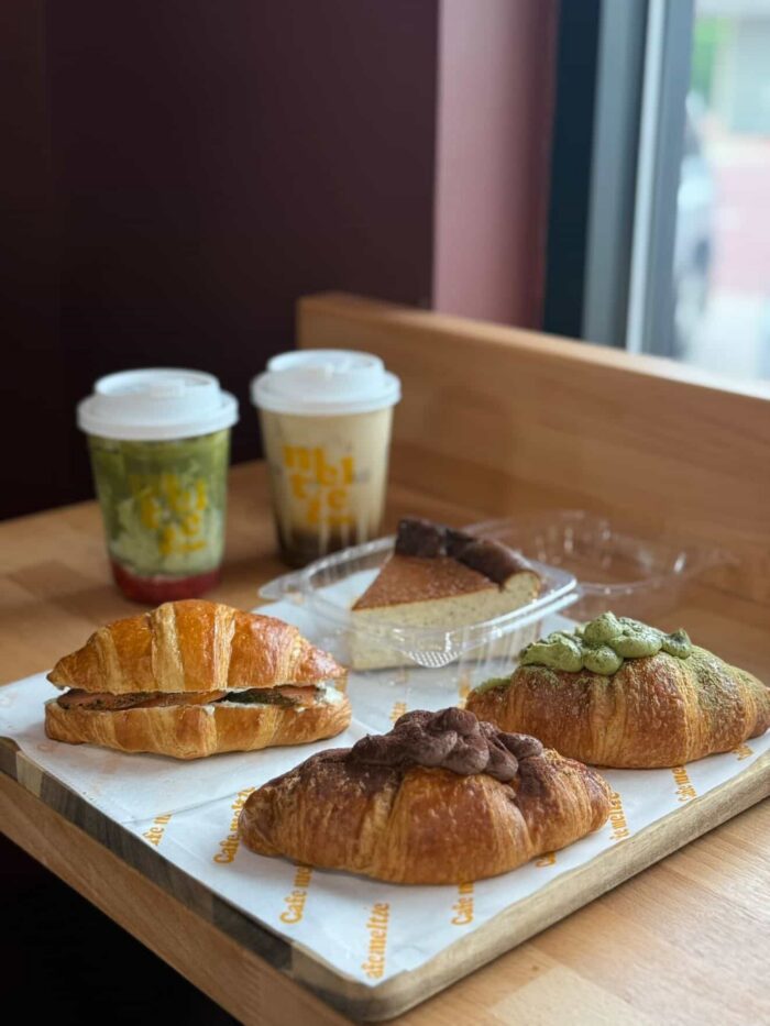 Three croissants with different toppings, a slice of cake, and two takeaway drinks on a wooden table by a window in a café.