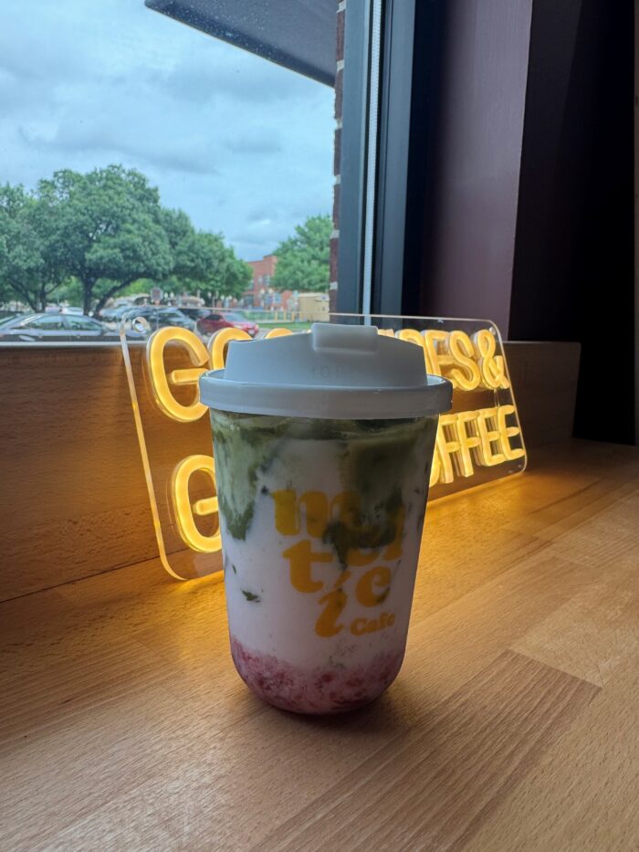 A colorful drink with green, white, and pink layers sits on a wooden table by a window, with a glowing yellow COOKIES & COFFEE sign and trees visible outside.