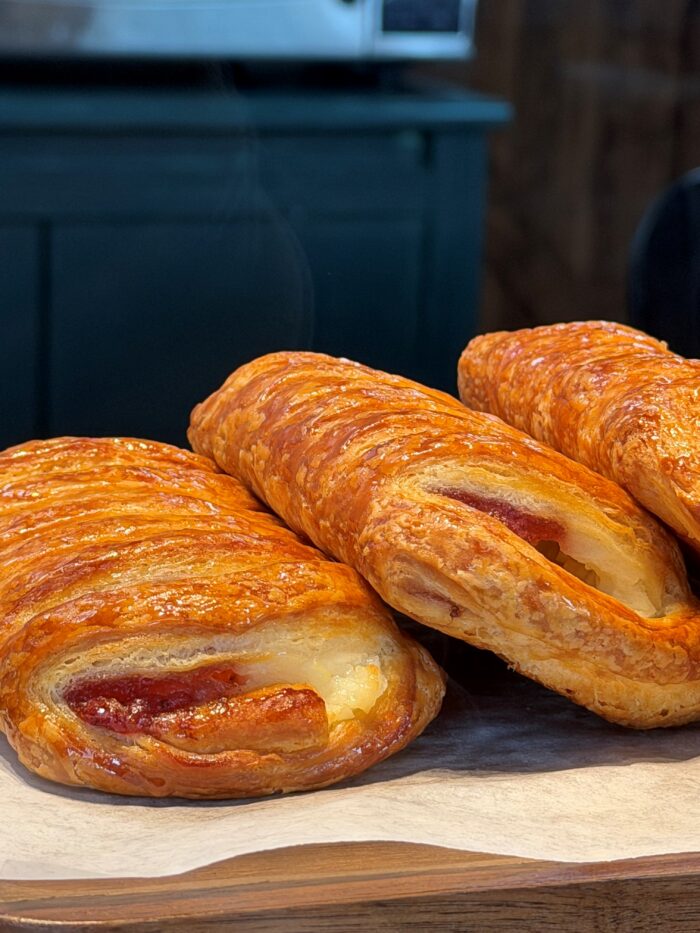 Three golden-brown, flaky pastries filled with cheese and what appears to be sausage, resting on parchment paper atop a wooden surface. A dark cabinet is blurred in the background.