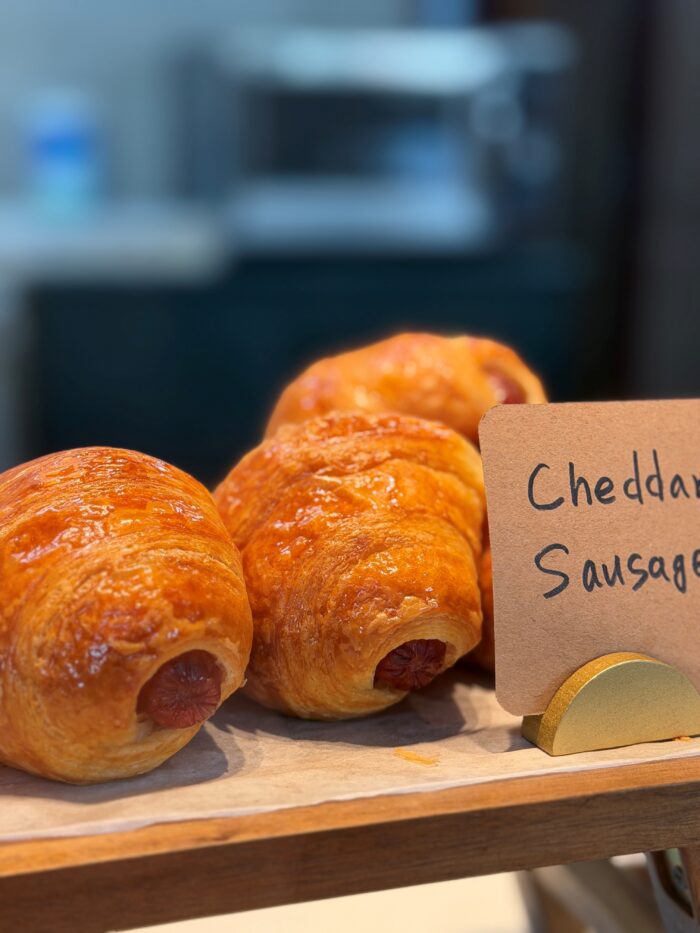 Three golden-brown cheddar sausage pastries are displayed on parchment paper, with a handwritten sign partially reading “Cheddar Sausage” in front of them. The background is softly blurred.