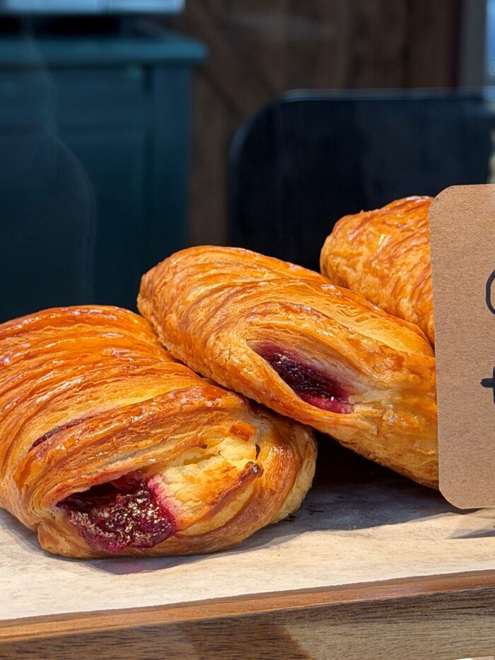 Three golden, flaky pastries filled with berry jam are displayed on a wooden tray, with a partial brown sign on the right side of the image.