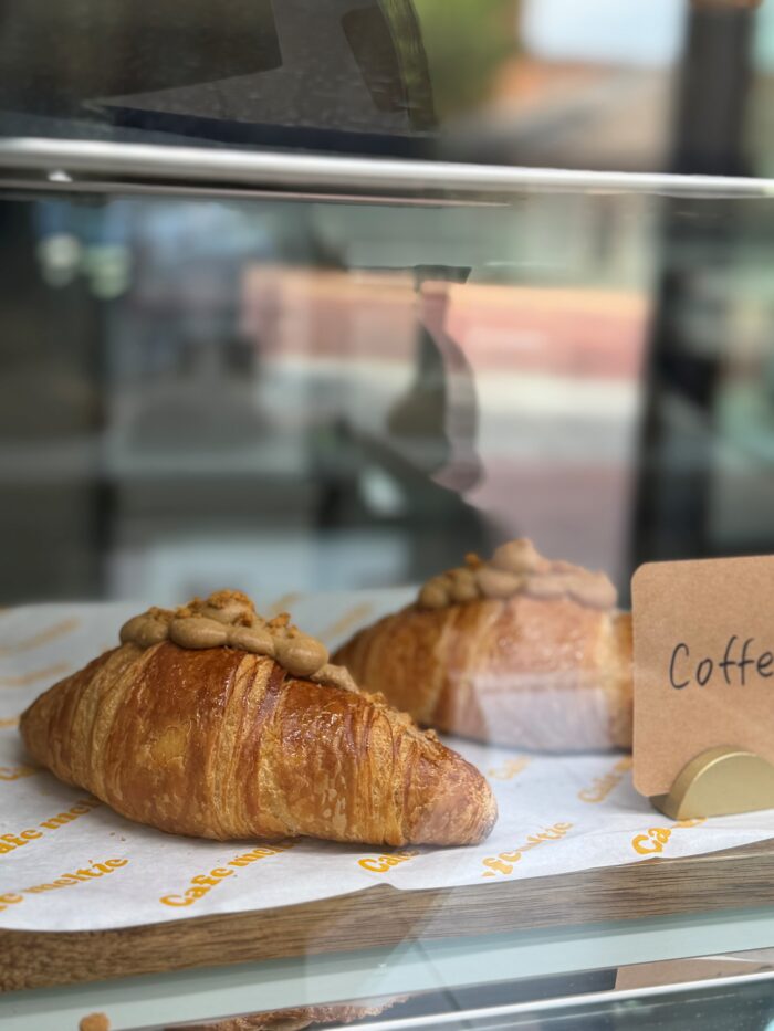 Two croissants topped with a dollop of cream are displayed on café-branded paper inside a glass case. A partial sign with the word Coffee is visible beside them.