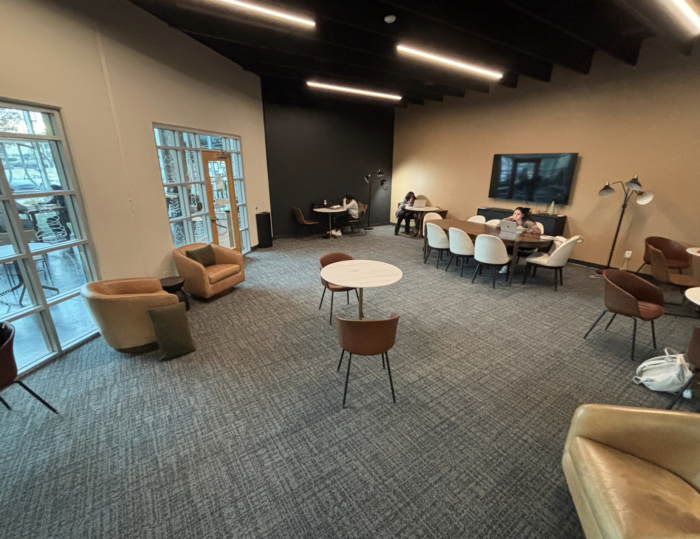 A modern lounge with several tables and chairs, some people working at a long table, large windows on the left, and neutral-colored walls and furniture. The space is well-lit with overhead lights.