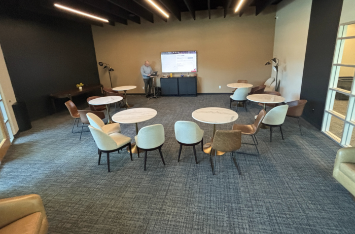 A conference room with round tables and chairs arranged in a semi-circle facing a large screen. A person stands near the screen at the front, presenting. The room has modern decor and overhead lighting.