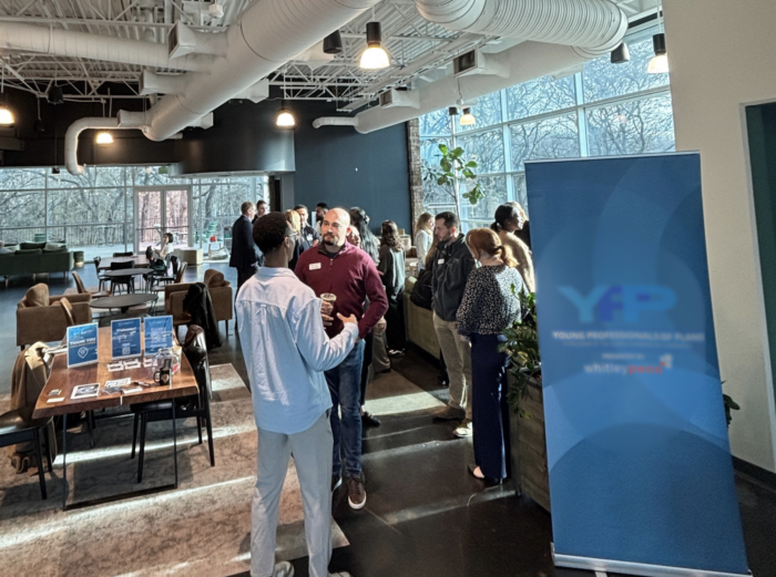 A group of people converse at an indoor networking event near large windows. A blue YP Young Professionals banner and information tables are visible. The space is modern with exposed ceilings and natural light.