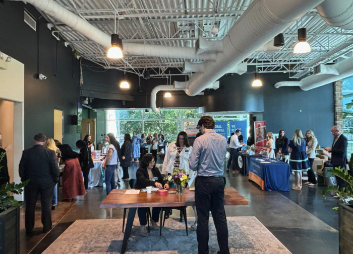 People mingle at a networking event in a modern, spacious room with tables, informational booths, and large windows. Some are seated at a table in the foreground while others interact in small groups around the room.