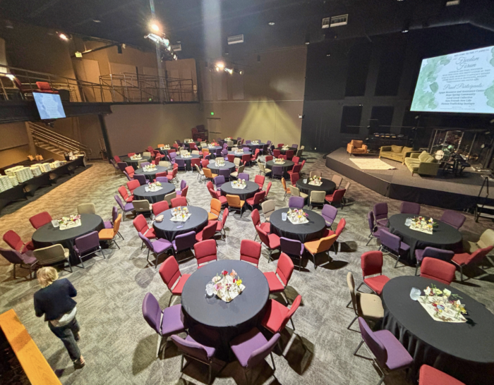 A large event space with round tables covered in black cloths, each set with flowers and surrounded by colorful chairs. A woman stands near the front, and a projection screen displays event information in the background.