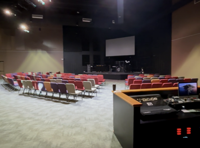 A modern church auditorium with rows of multicolored chairs facing a stage with instruments and a large screen; a sound booth with equipment is visible in the foreground.