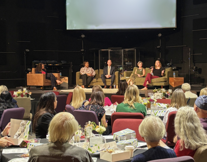 A panel of five people sits on stage having a discussion in front of an audience at a formal indoor event. Audience members sit at decorated tables with boxed meals and floral centerpieces.