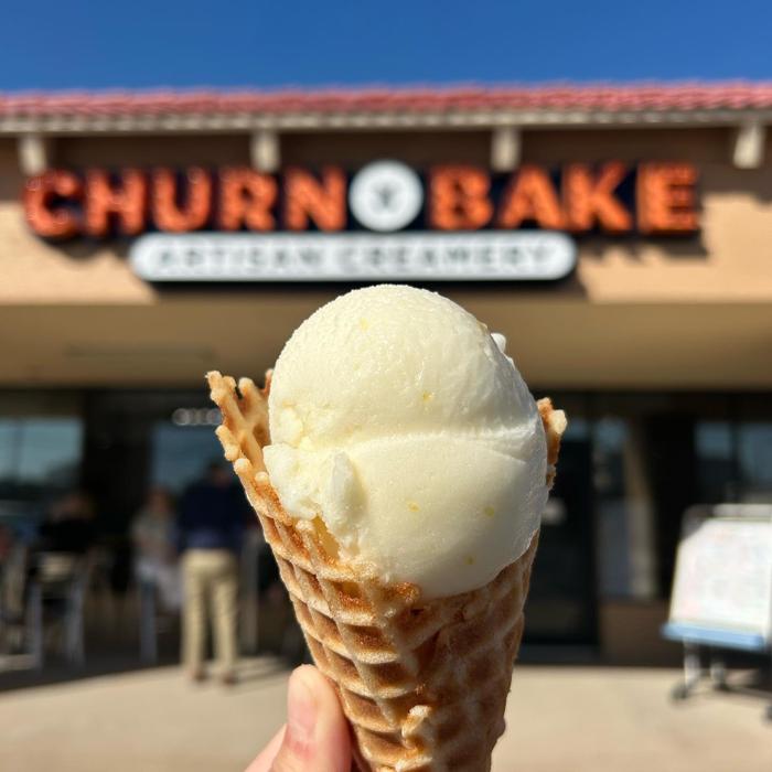 A hand holds an ice cream-filled waffle cone in front of a building with a sign that reads CHURN BAKE in large orange letters. The sky above is clear and blue.