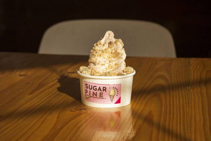 A cup of soft-serve ice cream topped with crushed crumbs sits on a wooden table. The Sugar Pine cup, featuring an ice cream cone graphic, is set against a dark background with a gray chair behind it.