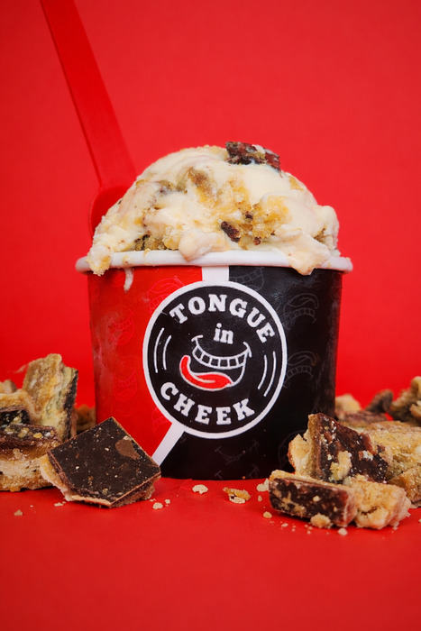 A cup of ice cream with chocolate chunks sits on a red surface, surrounded by more chocolate pieces. The black cup reads Tongue in Cheek and has a red spoon sticking out, all set against a bright red background.