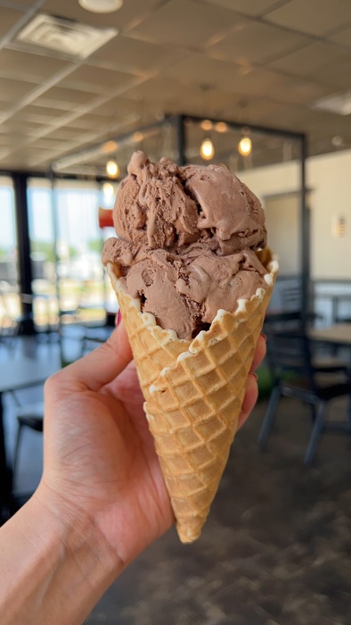 A hand holds a large waffle cone with two scoops of chocolate ice cream inside a bright, modern ice cream shop with tables and chairs in the background.