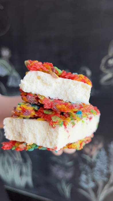 A hand holds two ice cream sandwiches with colorful, fruity cereal as the outer layers and thick vanilla ice cream in the middle, against a blurred chalkboard background.