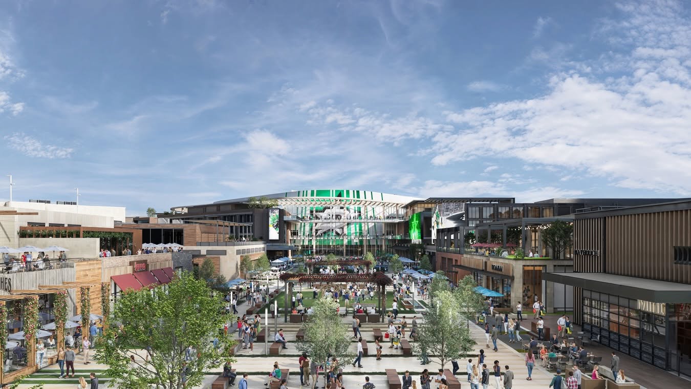 An outdoor shopping and dining plaza bustling with people under a partly cloudy sky, surrounded by modern buildings, greenery, and seating areas, with a large open area in the center.