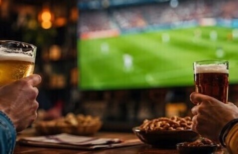 Zwei Personen mit Biergläsern in der Hand verfolgen in einer gemütlichen Bar ein Fußballspiel auf einem großen Fernsehbildschirm; vor ihnen stehen Schüsseln mit Snacks auf dem Holztisch.
