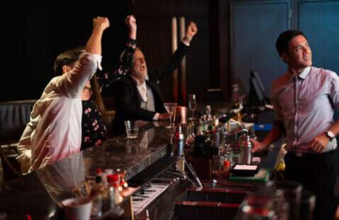Die Gäste in einer Bar feiern mit erhobenen Armen, während der Barkeeper lächelt und wegschaut.