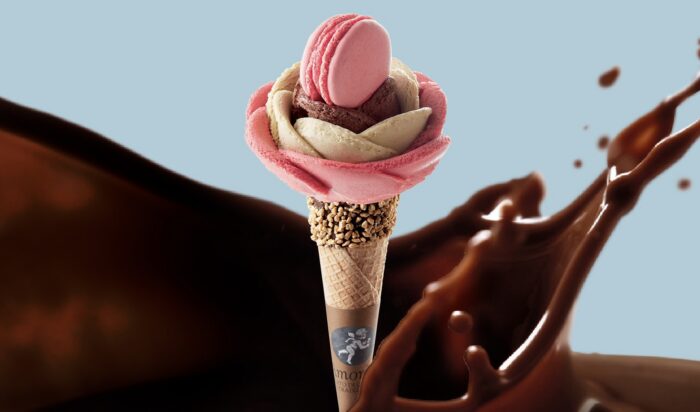A flower-shaped ice cream cone with pink and white petals, topped with a pink macaron, against a backdrop of swirling liquid chocolate. The cone is sprinkled with nuts near the top.