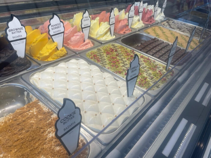 A display case filled with various flavors of gelato, each labeled with signs such as brownie batter, mango sorbet, pink guava, vanilla bean, and cookie crostata. The gelato is arranged in colorful, swirled mounds.