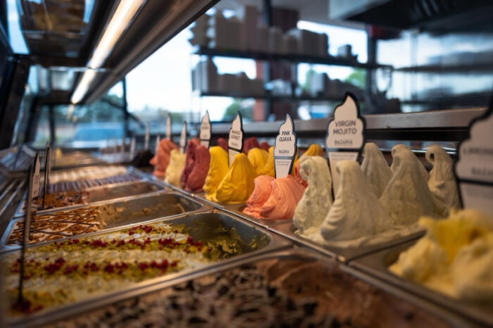 A display case filled with various colorful gelato flavors in metal trays, each labeled with signs. The gelato is arranged in swirled peaks and the background shows shelves and a window.